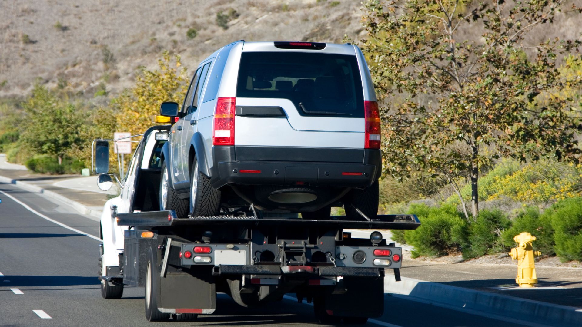 White SUV being towed on a flatbed truck along a roadside with trees