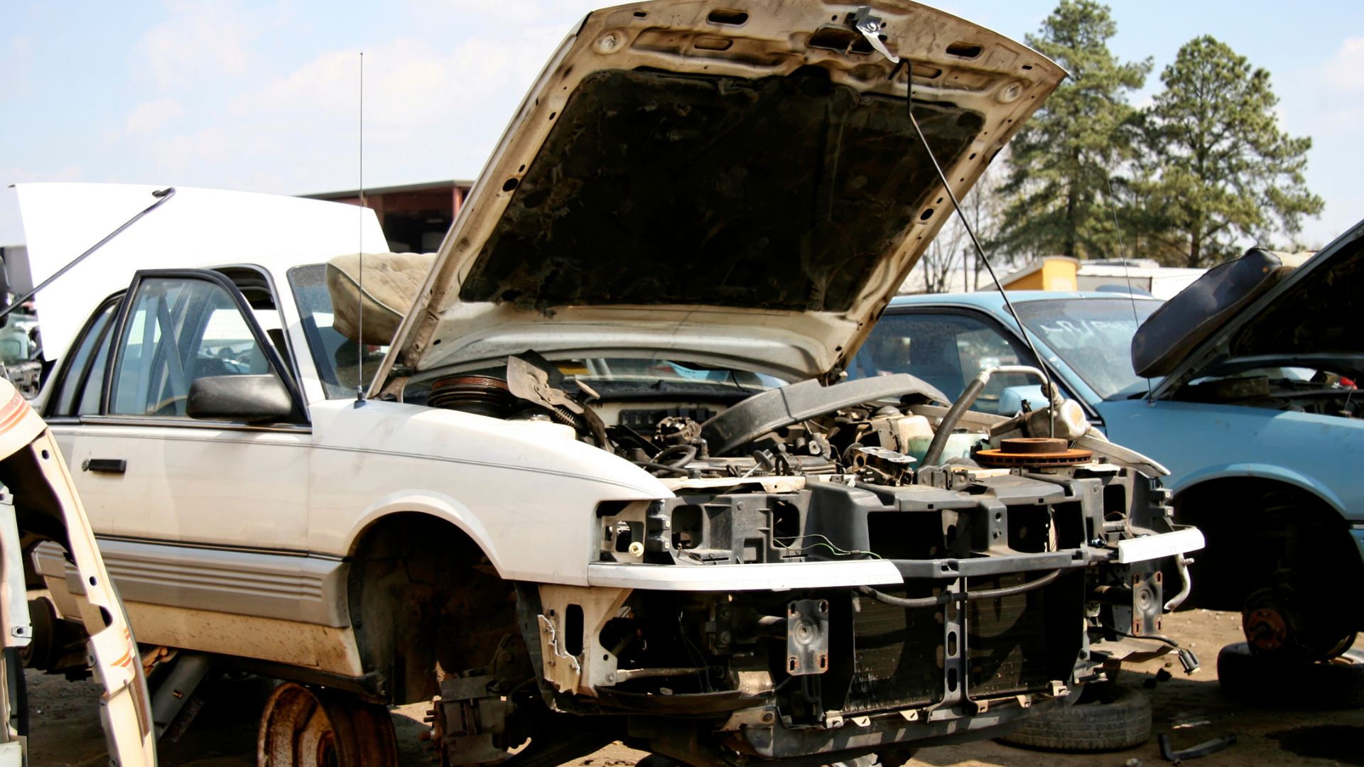 Damaged white car with open hood in junkyard, missing wheels and front panels
