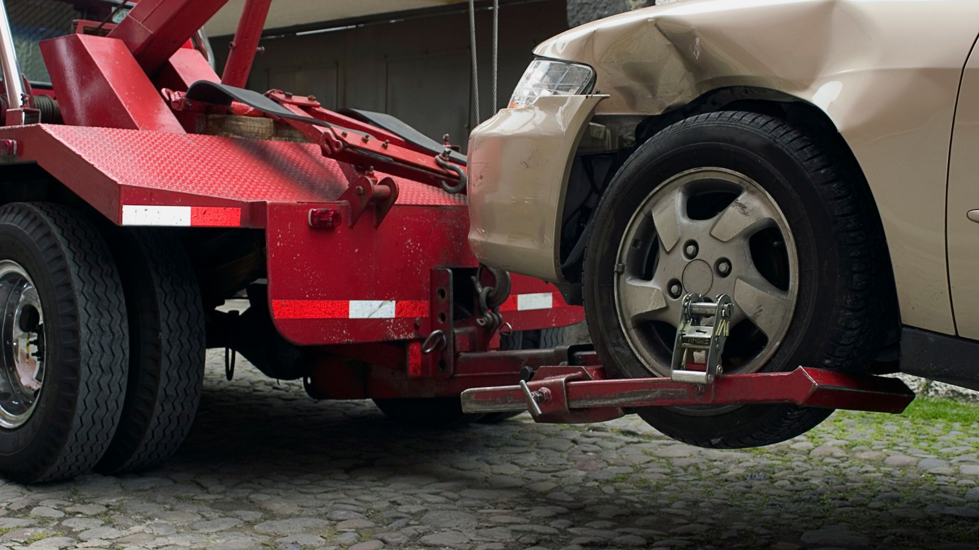 Red tow truck preparing to lift a beige car on cobblestone ground